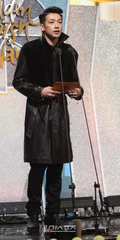 a man standing in front of a microphone on top of a stage wearing a black coat