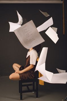 a woman is sitting on a chair with papers flying around her and paper birds in the air