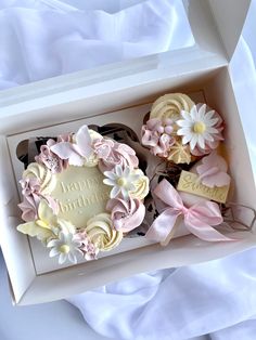 two cupcakes in a white box with pink and yellow flowers on them, one has a happy birthday sign