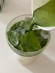 a person pouring green liquid into a glass filled with ice cubes and greens in it