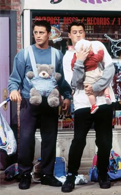 two men are standing on the sidewalk with a teddy bear in front of a store