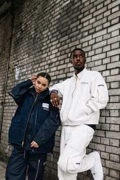 two men standing next to each other in front of a brick wall wearing white and blue clothing