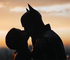 a man in a batman costume kissing a woman on the forehead at sunset with city lights behind him