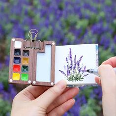 a person holding an art book with paint and watercolors in front of purple flowers