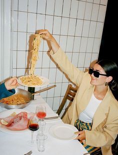 two women sitting at a table with plates of food in front of them and one woman holding up a piece of pasta