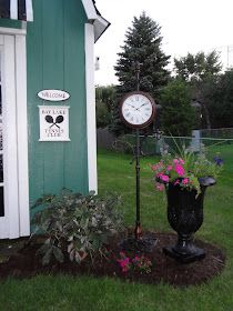 a clock sitting next to a potted plant in front of a green building with white trim