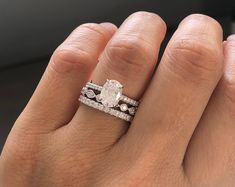 a woman's hand with two wedding rings on top of her finger and the ring has an oval shaped diamond surrounded by smaller round diamonds