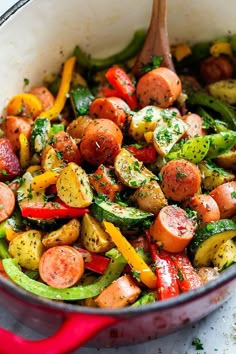 a pan filled with vegetables and sausages on top of a white countertop next to a wooden spoon