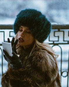 a woman wearing a fur hat and gloves looking at her tablet while standing outside in the snow