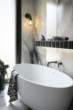 A stylish bathroom featuring a white freestanding tub, a modern wall light illuminating the space, and marble walls complemented by a sleek shelf displaying decorative items and essentials.