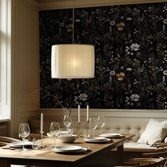 a dining room table set with place settings and wine glasses on it, in front of a floral wallpaper