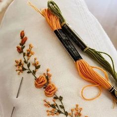 two needles and some orange flowers on a white table cloth with embroidery thread in the middle