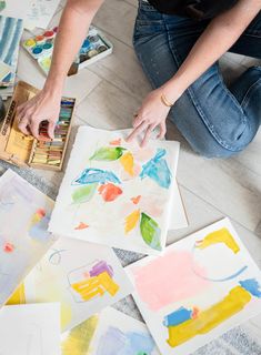 a person is painting on paper with watercolors and paintbrushes while sitting on the floor