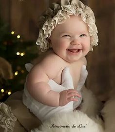 a smiling baby wearing a white dress sitting in front of a christmas tree