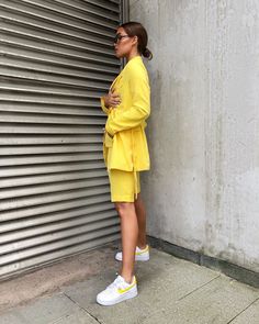 Fashion 2024 shines bright with this vibrant, sunshine-yellow ensemble, embodying the joyous spirit of spring and summer streetwear. The woman stands poised, her chic blazer and shorts set offering a playful yet polished look, cinched at the waist for a flattering silhouette. Paired with classic white sneakers that feature a pop of yellow, she presents a harmonious blend of casual comfort and style. Her sleek sunglasses and pulled-back hair accentuate the clean lines and fresh vibe of the outfit. This is a statement of bold color and relaxed tailoring, showcasing how Outfits 2024 celebrates personal style with energetic hues and effortless grace. Blazer Shorts Outfit, Athleisure Trend, Chic Blazer, Summer Streetwear, The Embrace, Classic Suit, Blazer And Shorts, Woman Standing