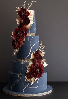 a three tiered blue cake with red flowers on it's side and gold accents