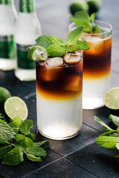 two glasses filled with ice and mint on top of a table next to limes