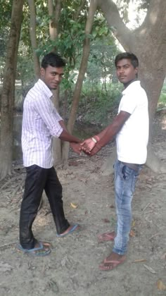 two men holding hands in front of trees and dirt ground with one man wearing sandals