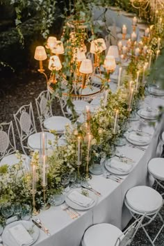 an outdoor dinner table with candles, flowers and greenery on the tables is surrounded by white chairs