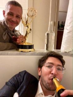 two men sitting at a table and one drinking from a wine bottle while the other is holding an award