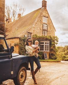 a woman holding a baby standing in front of a blue truck with a house behind her