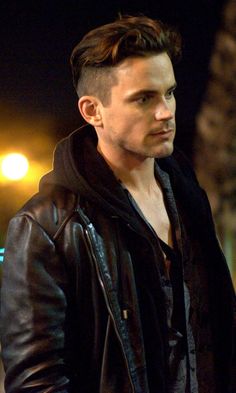 a young man in a leather jacket looks off into the distance at night with street lights behind him