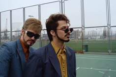 two men in suits and sunglasses standing on a tennis court with a fence behind them