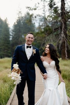 a bride and groom holding hands while walking down a wooden walkway in the woods with tall grass