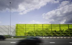 a car driving down the road past a large building with yellow panels on it's side