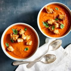 two bowls of tomato soup with bread croutons and basil leaves on the side