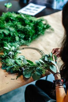 a woman is making a wreath out of greenery on a wooden table with scissors