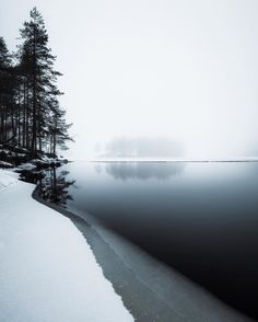 a body of water surrounded by snow covered ground and trees in the distance with fog