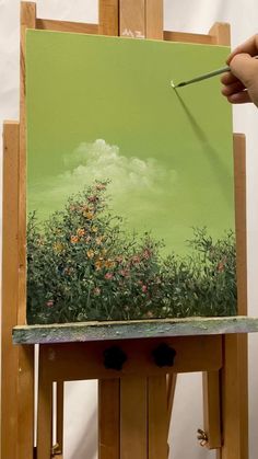 someone is painting flowers on an easel with a green background and white clouds in the sky