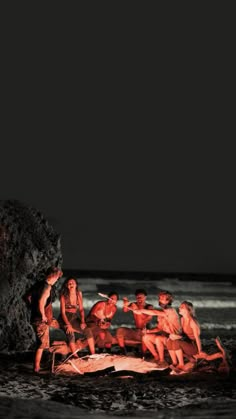 group of people sitting around an open fire pit on the beach at night with dark sky in background