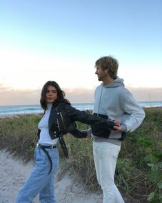 a man and woman walking on the beach next to each other with their hands in their pockets