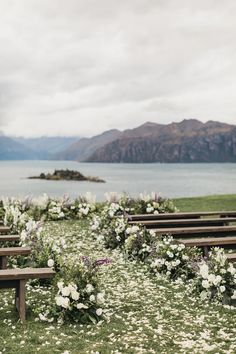 an outdoor ceremony setup with flowers and benches on the grass by the water in front of mountains