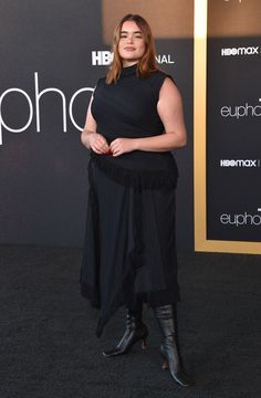 a woman standing in front of a black background wearing a long skirt and high heeled boots