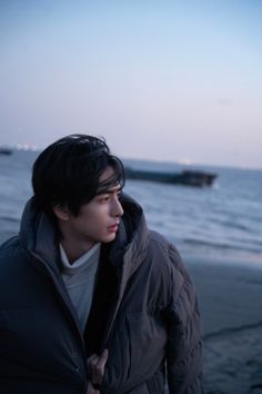 a man standing on top of a beach next to the ocean at dusk with his hands in his pockets