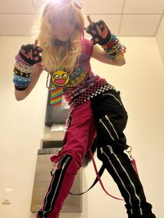 a woman in pink pants and colorful shirt holding two hands up to her head while standing next to a wall