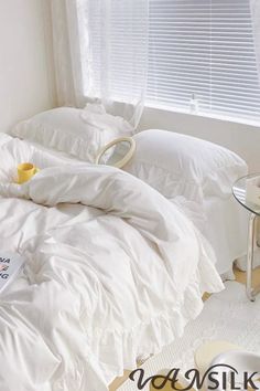 an unmade bed with white sheets and pillows in front of a small table next to a window