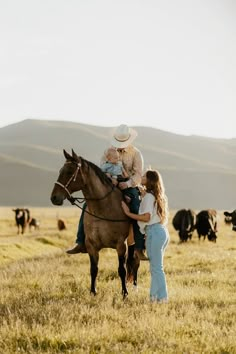 a woman holding a child on the back of a horse in a field full of cows