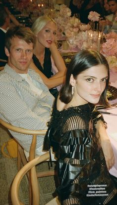 two men and a woman sitting at a table with flowers in the background, all dressed up