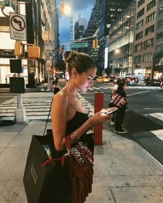 a woman is standing on the sidewalk looking at her cell phone and holding a bag