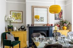 a dining room table and chairs with yellow lamps hanging over it's head, in front of a fireplace