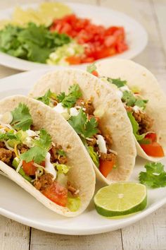 three tacos on a plate with limes and tomatoes