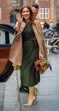 a woman in a green dress and coat waves to the camera while standing next to a car