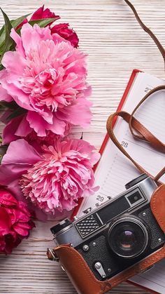 a camera, notebook and pink flowers on a wooden table with a brown leather case