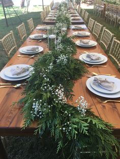 a long wooden table with plates and place settings on it, surrounded by greenery