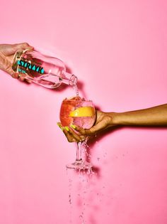 two hands are holding an apple and pouring it into a wine glass on a pink background