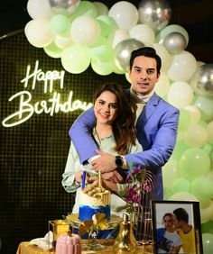 a man and woman standing in front of a birthday cake with candles on it, surrounded by balloons